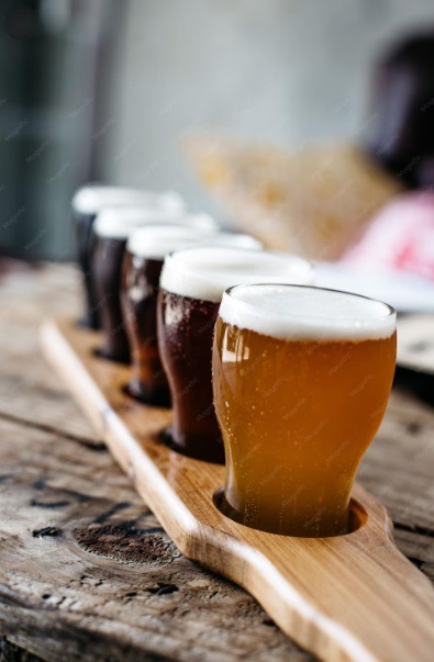 A wooden flight paddle holds six small glasses of craft beer, showcasing a variety of colors and styles, set on a rustic wooden table.