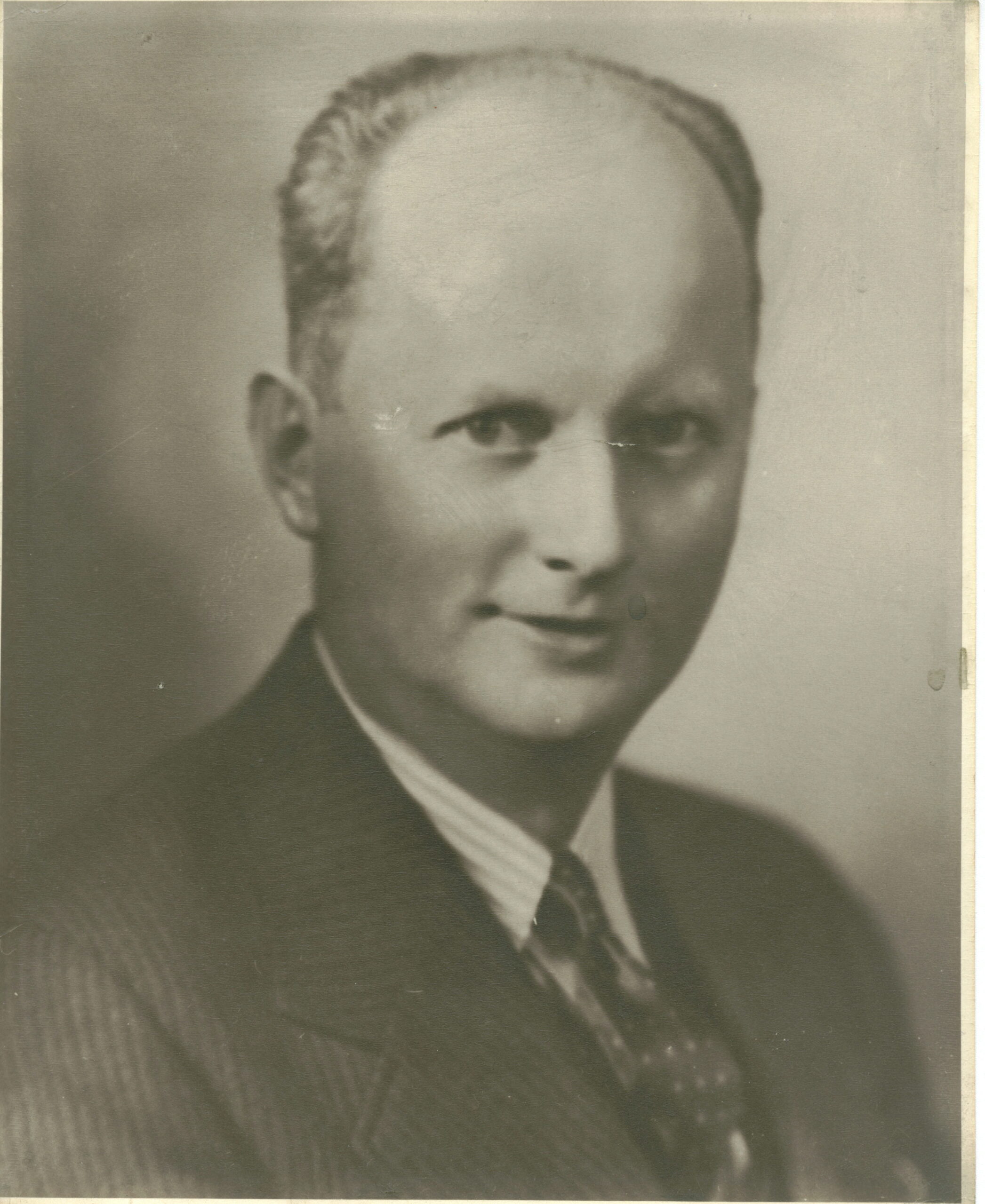 Black and white portrait of a man in a suit, smiling slightly. His hair is neatly combed, and the background is softly blurred, emphasizing his face.