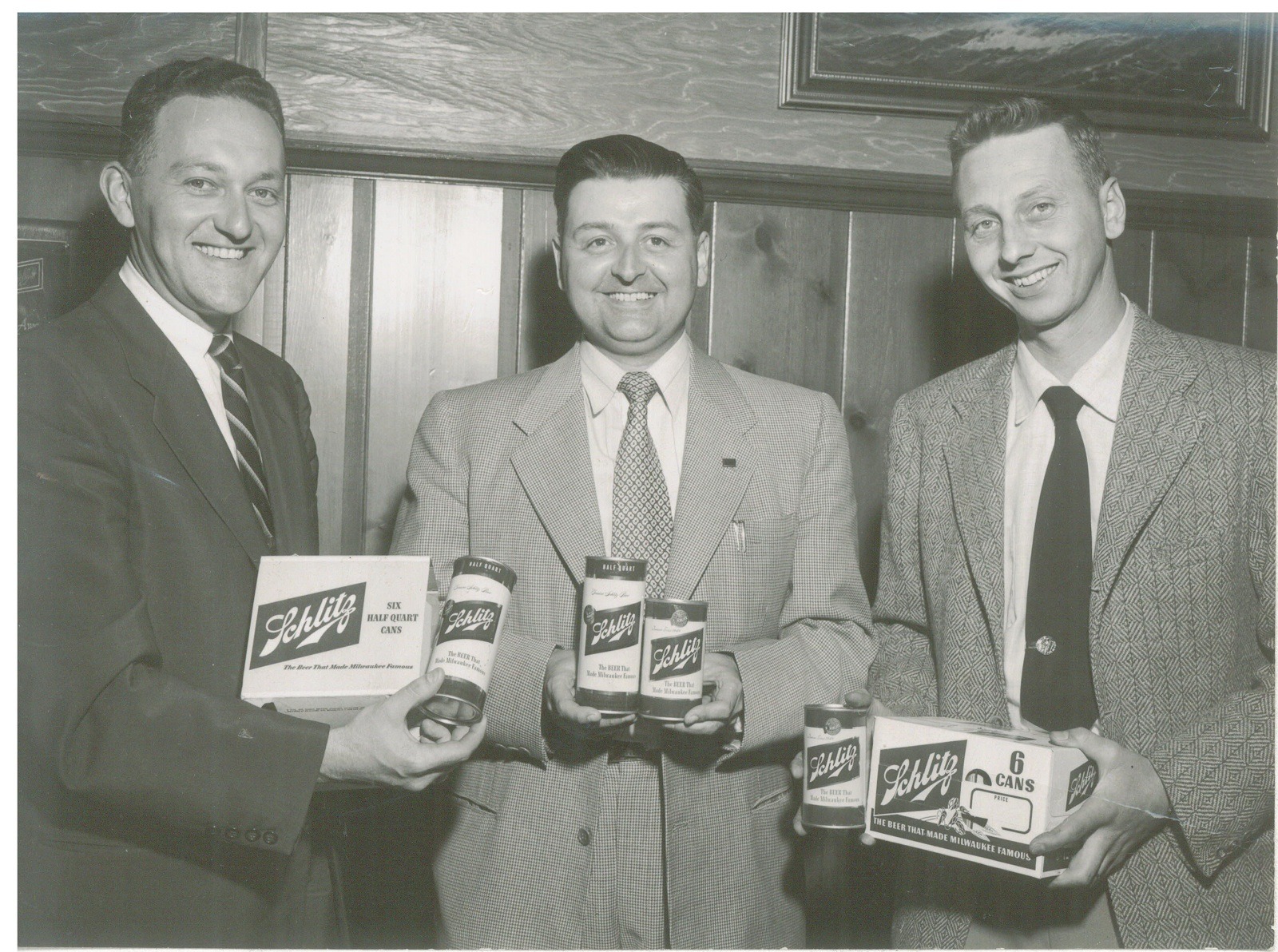 Three men display Schlitz beer products, including cans and a box, against a wooden backdrop. The photo highlights their promotional involvement with the brand.