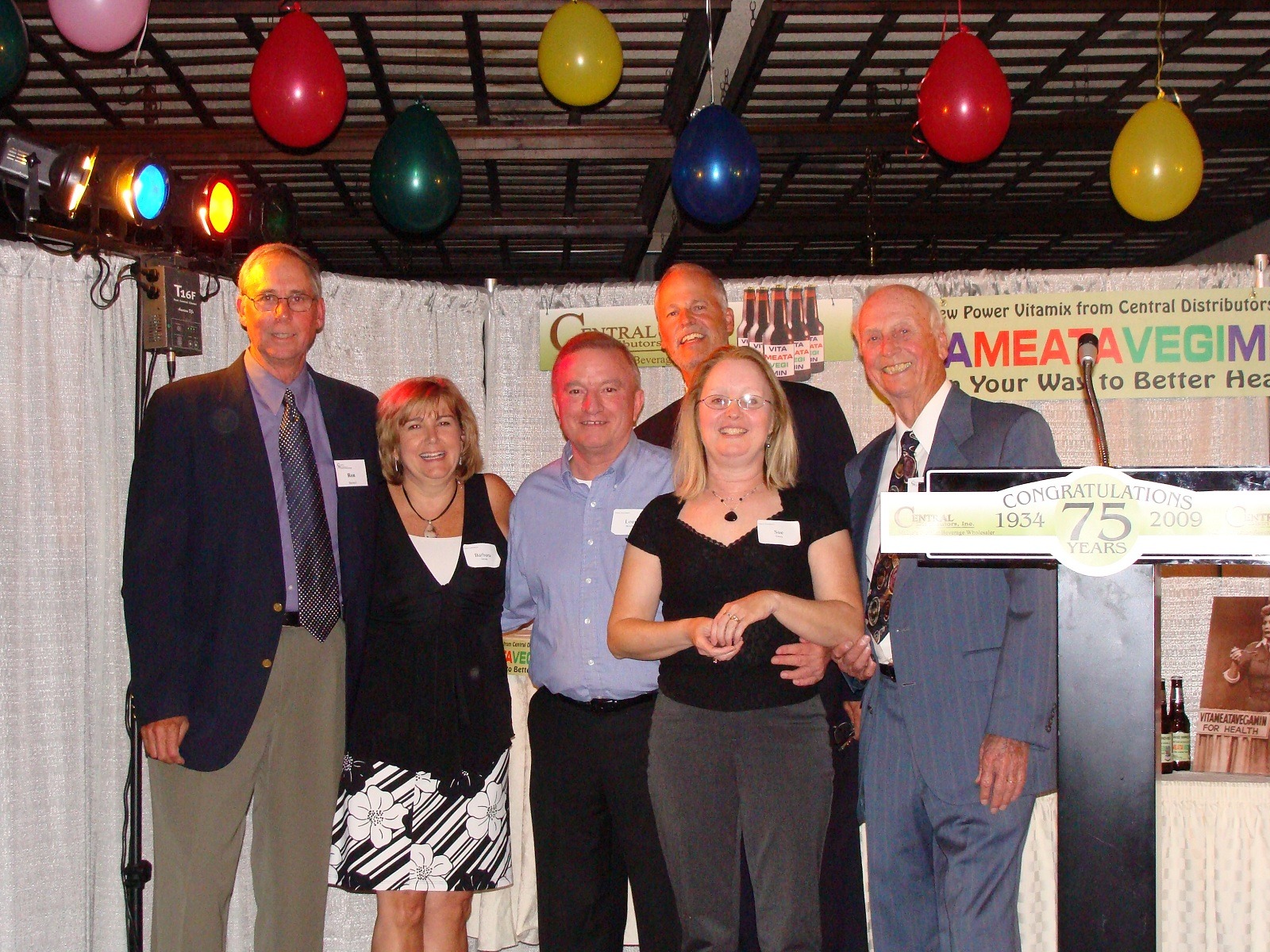 Group of six people celebrating a 75th anniversary, standing in front of a festive backdrop with balloons and a congratulatory sign.