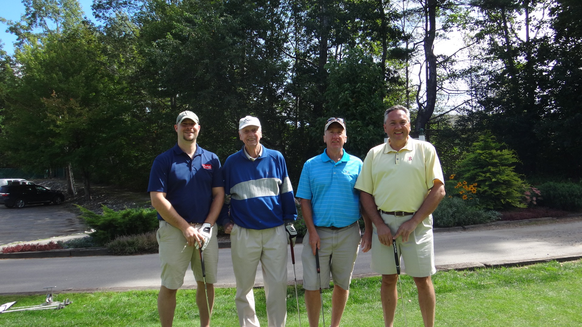 Four men stand together on a golf course, each holding a golf club. They are dressed in casual golf attire, smiling against a backdrop of trees.